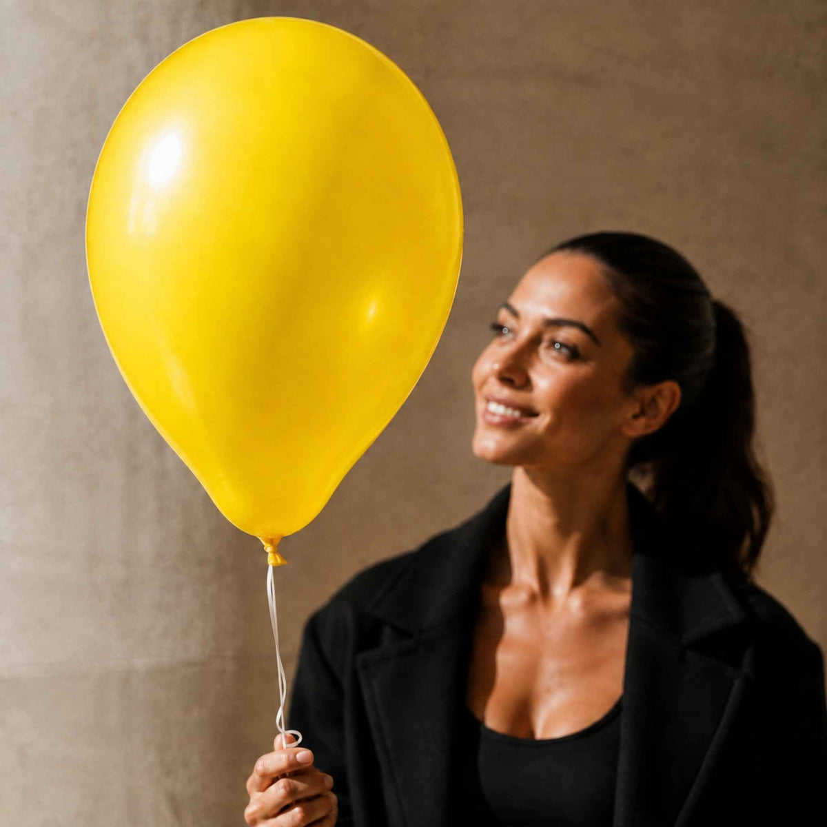 A woman in a black blazer holds a large, bright yellow balloon against a textured beige wall background.